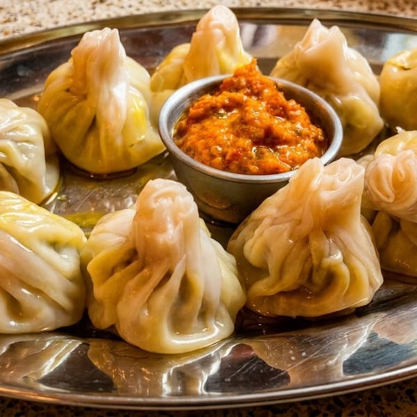 A silver platter holds a dozen steamed momos surrounding a small bowl of orange chili sauce.